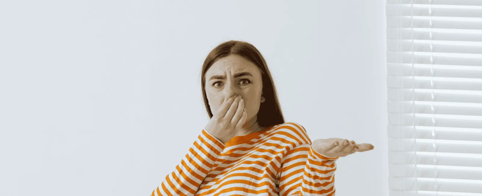 Young woman in orange striped shirt holding nose with disgust against white wall and window blinds