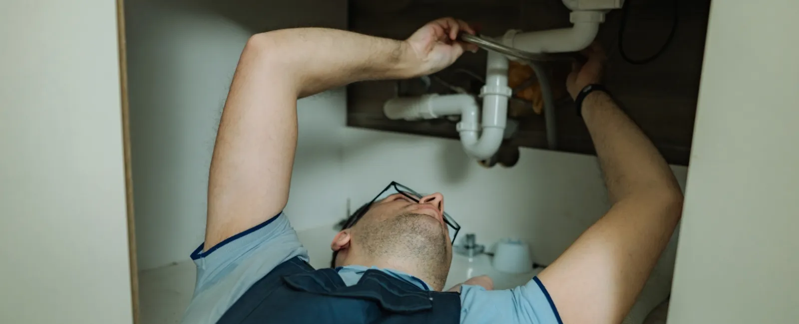 Plumber wearing glasses fixes white PVC pipes under a kitchen sink using a wrench in a modern cabinet.