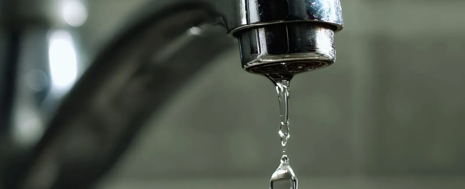 Close-up of a chrome faucet with a water droplet falling, blurred background of tiles.