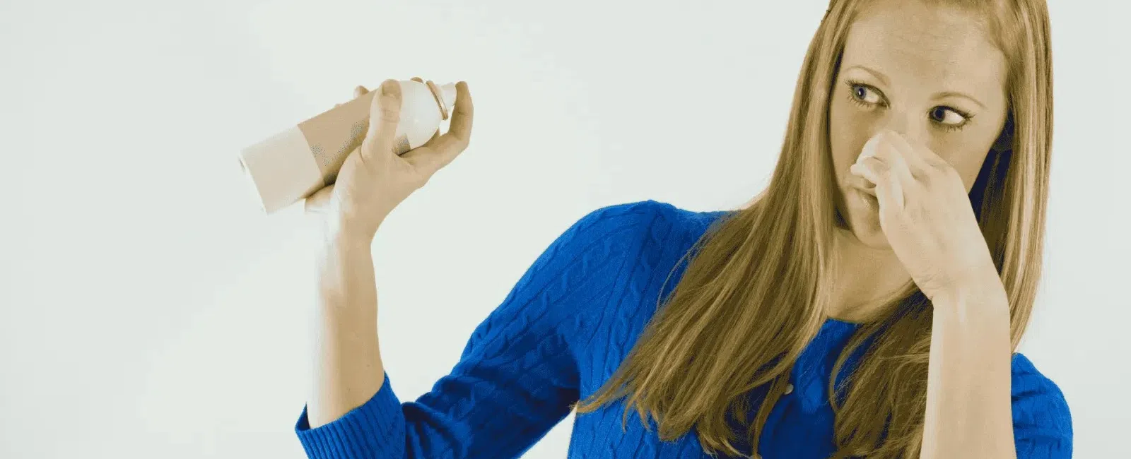 Woman in blue sweater pinching nose and shaking spray can to stop bad odor indoors.