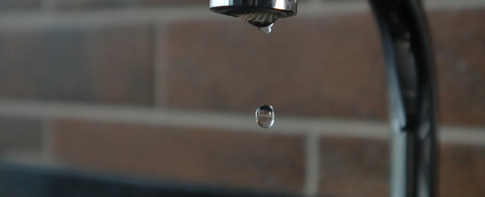 Close-up of a water droplet falling from a kitchen faucet with blurred tiled wall background