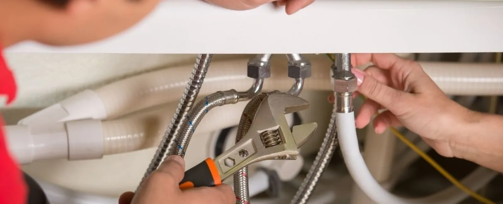 Plumber using a wrench to fix under-sink plumbing connections with flexible hoses and pipes.