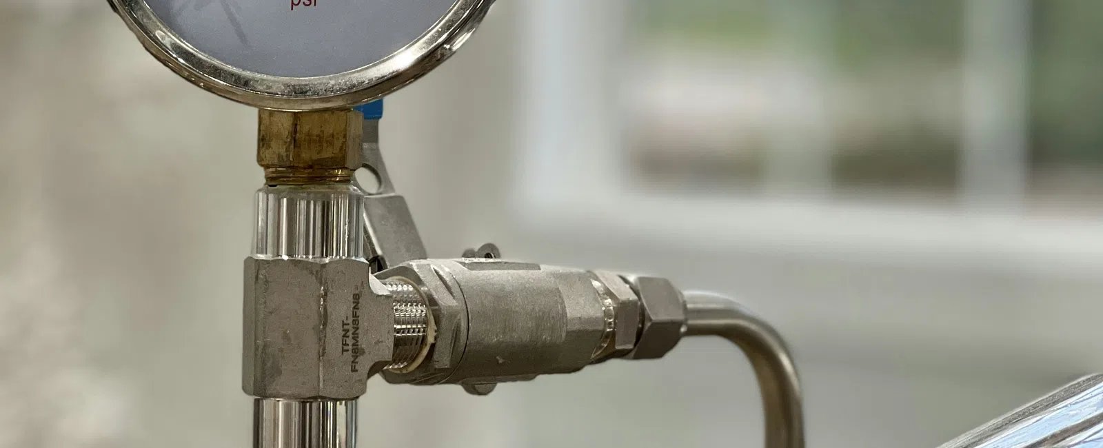 Close-up of a pressure gauge attached to polished metal piping system indoors near a window.