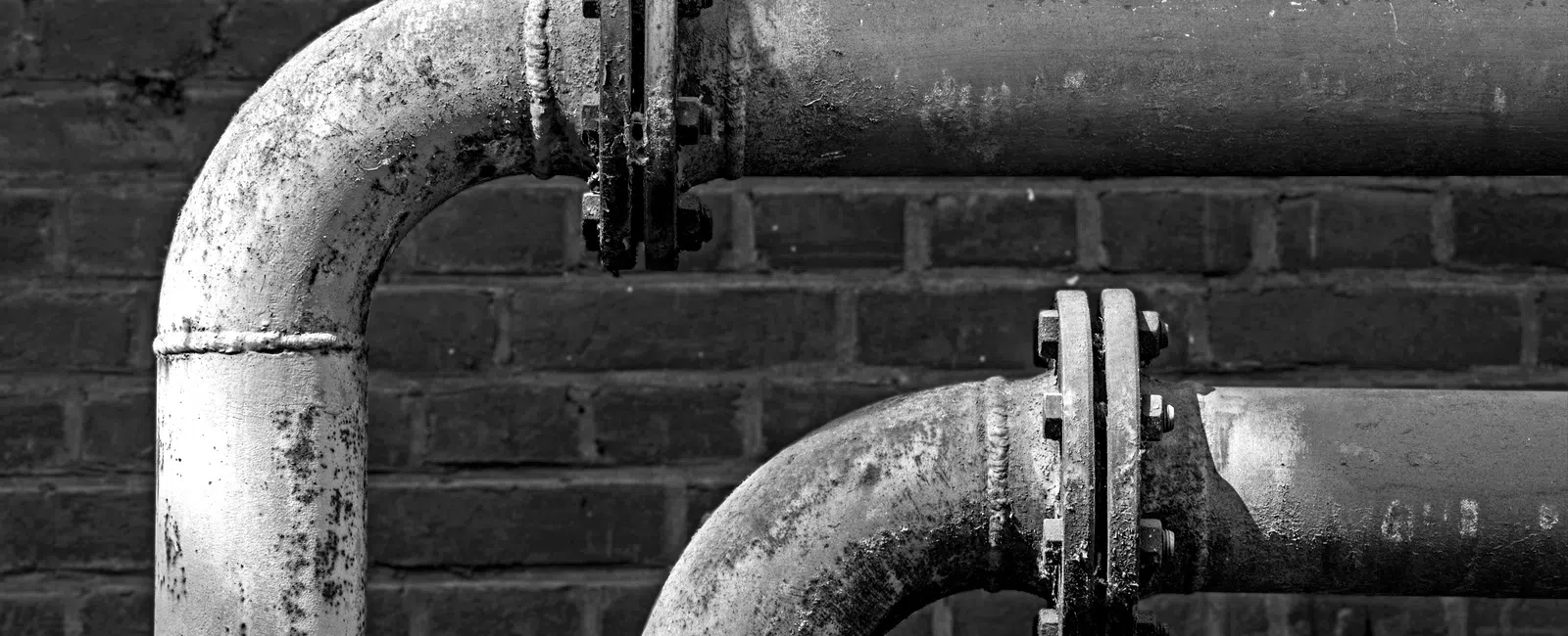 Close-up of two old metal pipes with flanges against a dark brick wall in black and white.