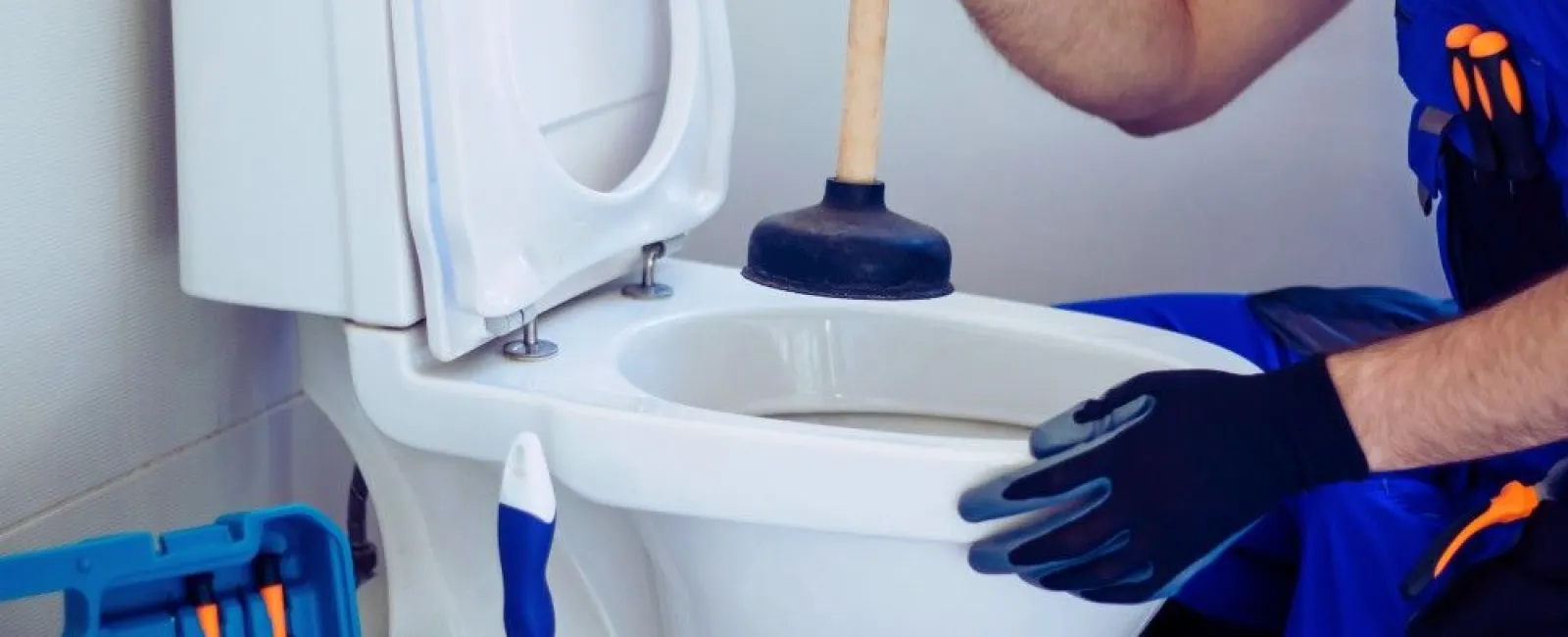 Plumber using a plunger to unclog a white toilet in a bathroom with toolkit nearby.