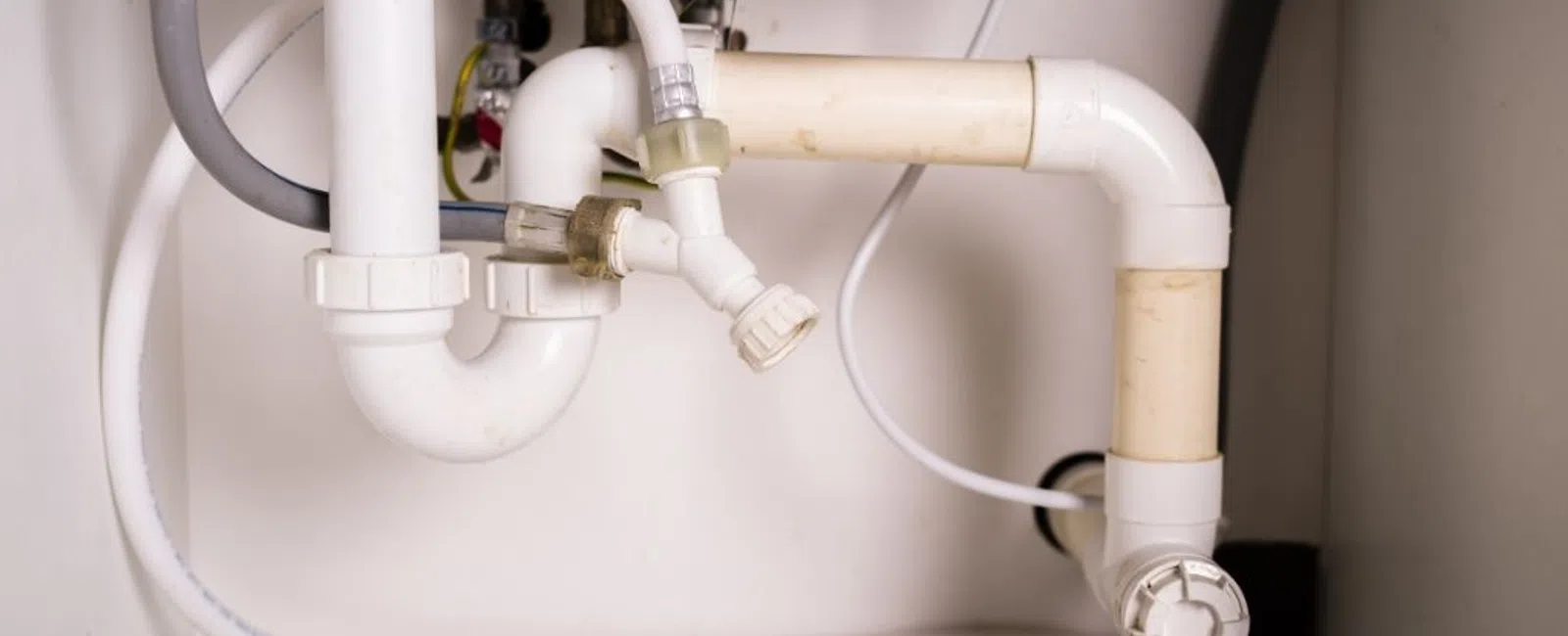 White plastic plumbing pipes with connectors and valves under a sink cabinet against a light background.
