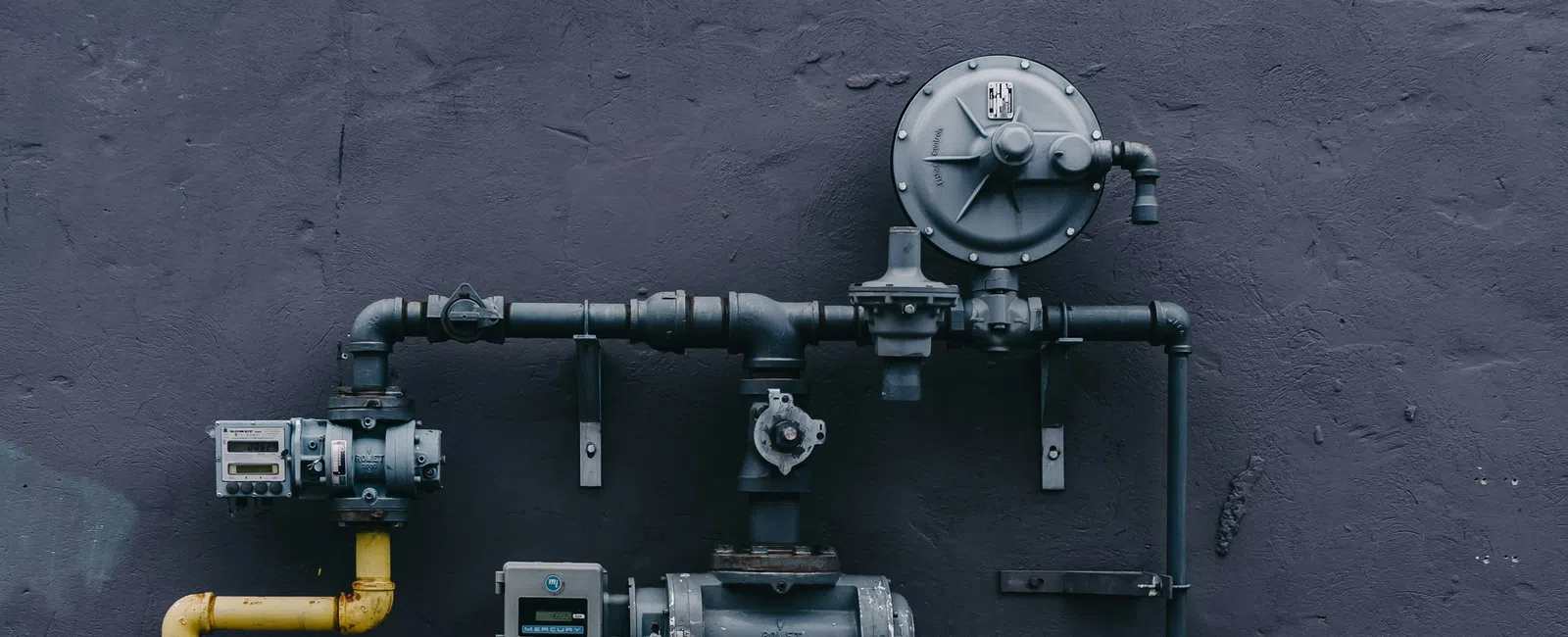 Gas meter and piping setup with yellow and black pipes mounted on a dark gray wall outdoors.