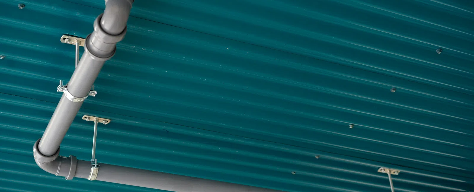Gray metal pipes with joints mounted on a teal corrugated metal ceiling with metal brackets.