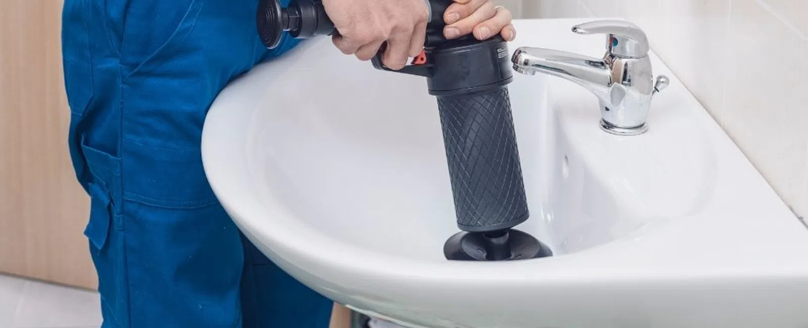 Plumber in blue overalls using a manual drain auger to unclog a white bathroom sink drain effectively.