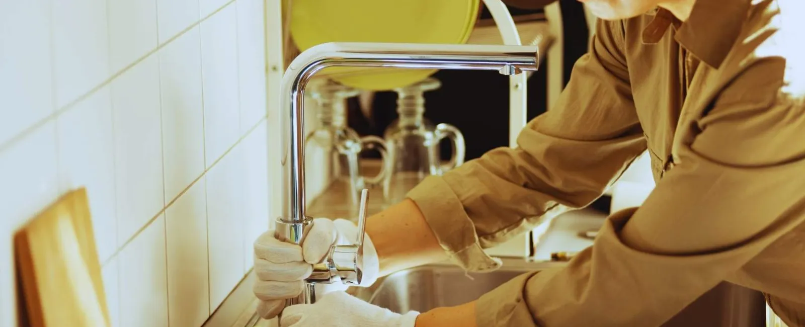Person wearing gloves fixing a modern kitchen faucet over a stainless steel sink with dishware in the background