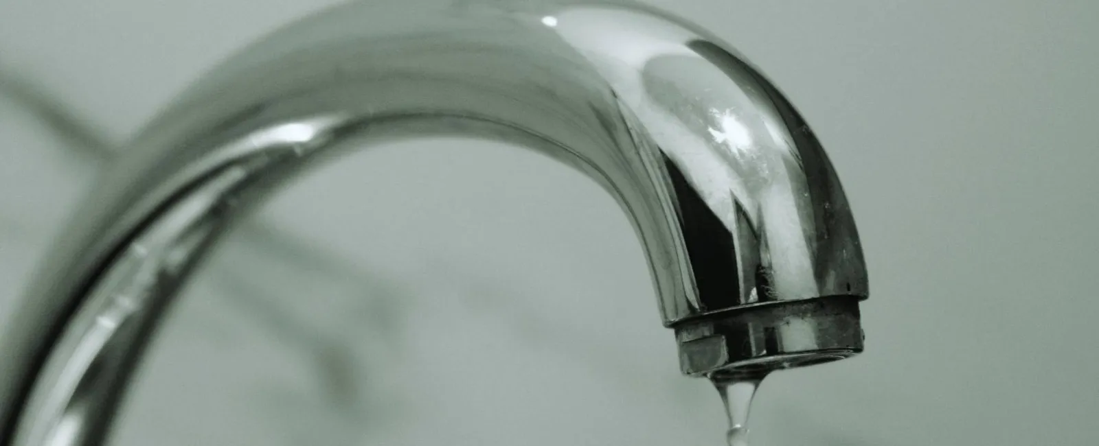 Close-up of a shiny chrome faucet with a water droplet falling against a blurred background.
