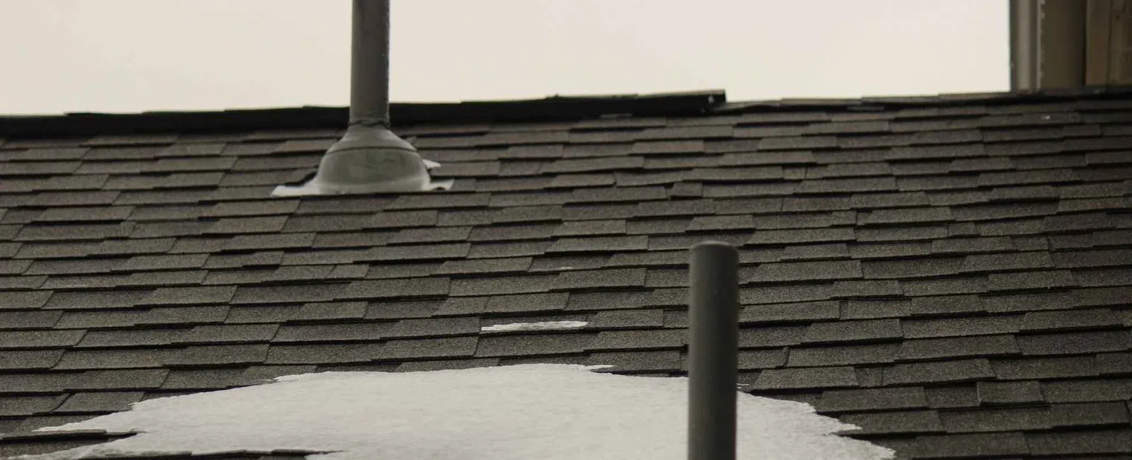 Snow partially covering a dark asphalt shingle roof with two black vent pipes against a white sky background.
