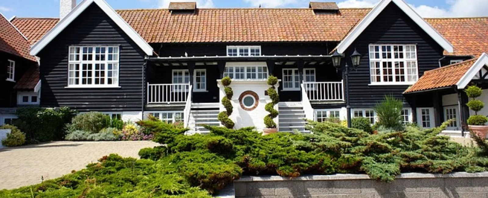 Large black and white house with red tile roof surrounded by green shrubbery under a blue sky with clouds.
