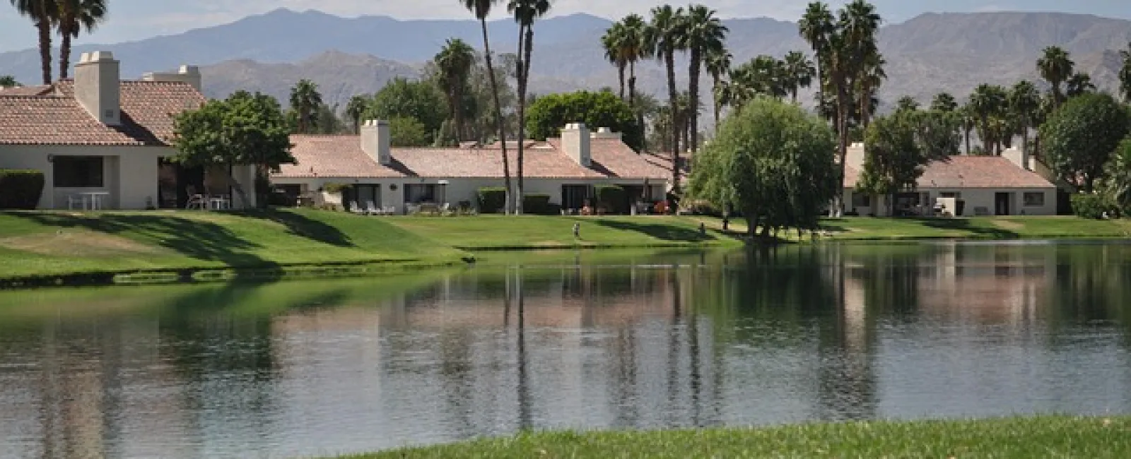 Lakeside view of suburban homes with palm trees and mountains under a partly cloudy sky.