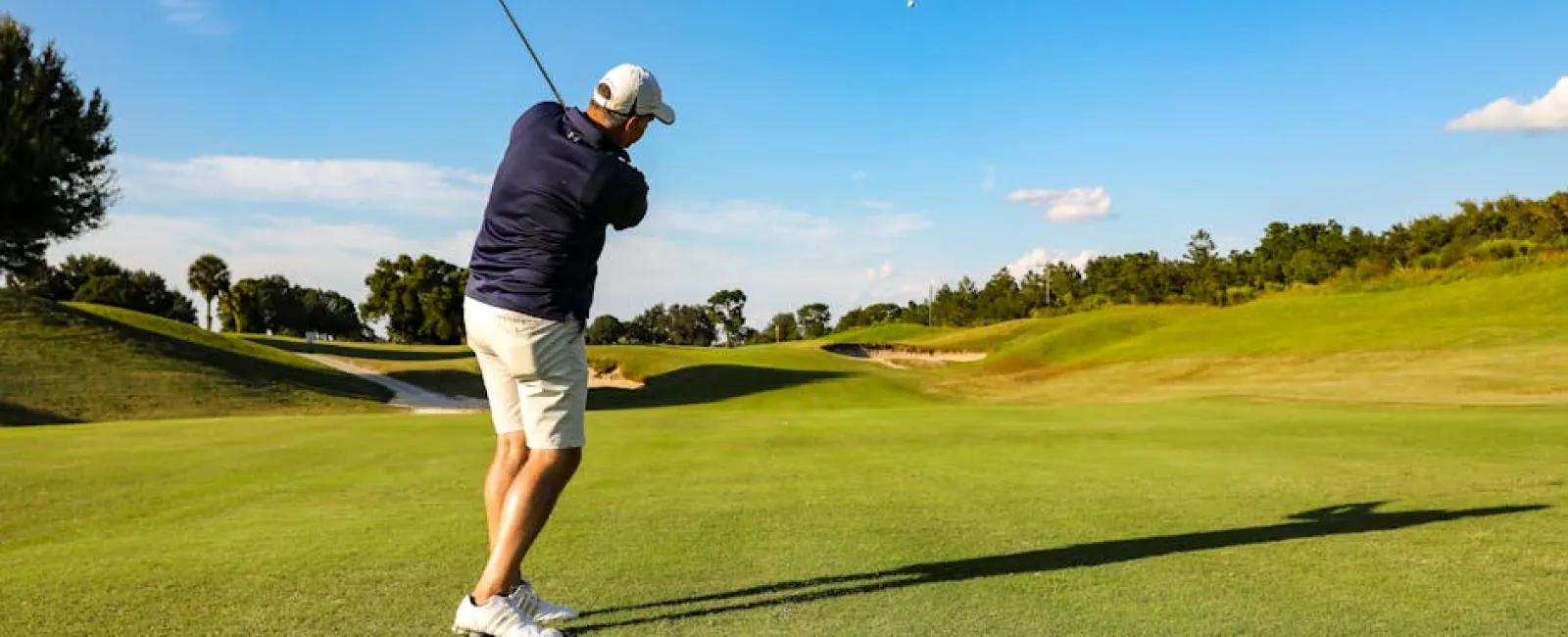 Golfer in navy jacket and white shorts hitting a shot on a sunny green golf course with blue sky.