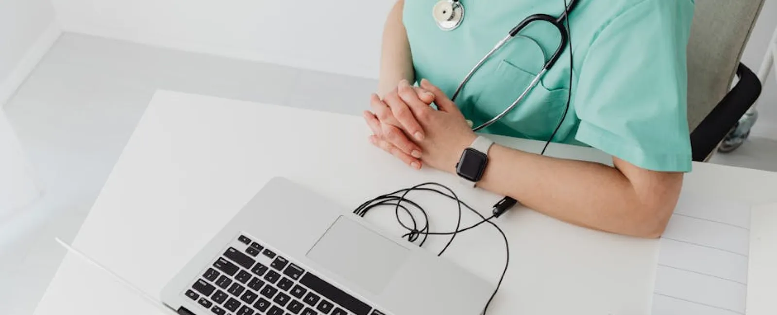 Healthcare professional in green scrubs with stethoscope, headset, and laptop at a white desk, hands clasped.