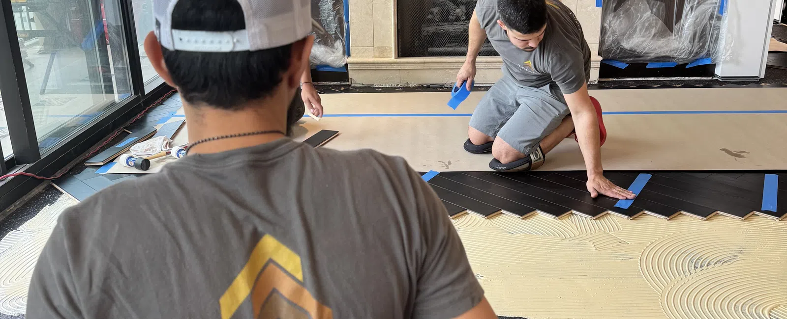 Two workers installing dark hardwood flooring in a modern living room with a fireplace and large windows.