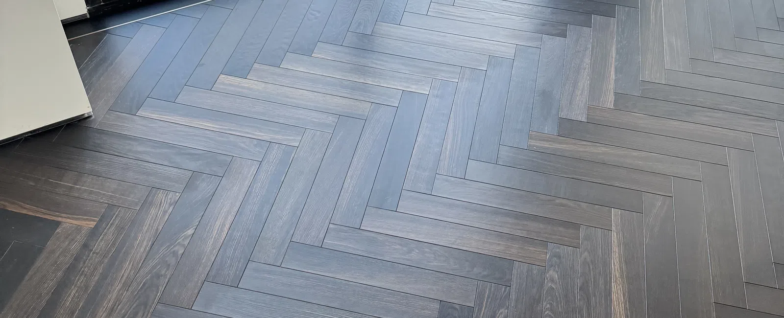 Dark wood herringbone patterned floor in a modern room with large floor-to-ceiling window overlooking autumn trees.