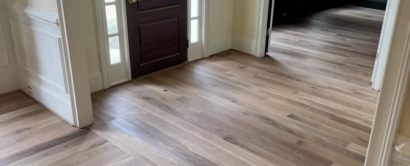 Entrance hallway with dark wooden front door, large windows, and light hardwood flooring in a bright home.