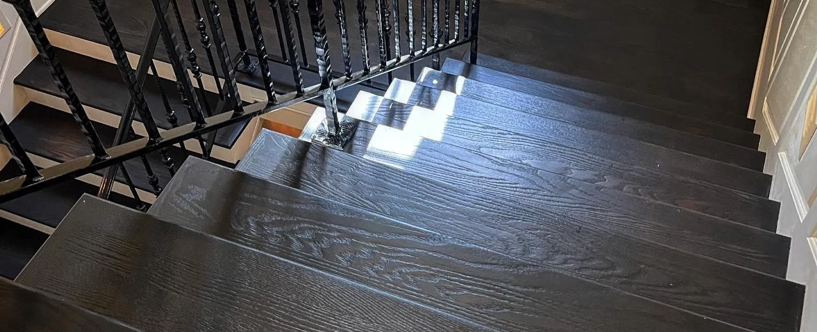 Dark wooden staircase with black wrought iron railing and white paneled walls in bright natural light