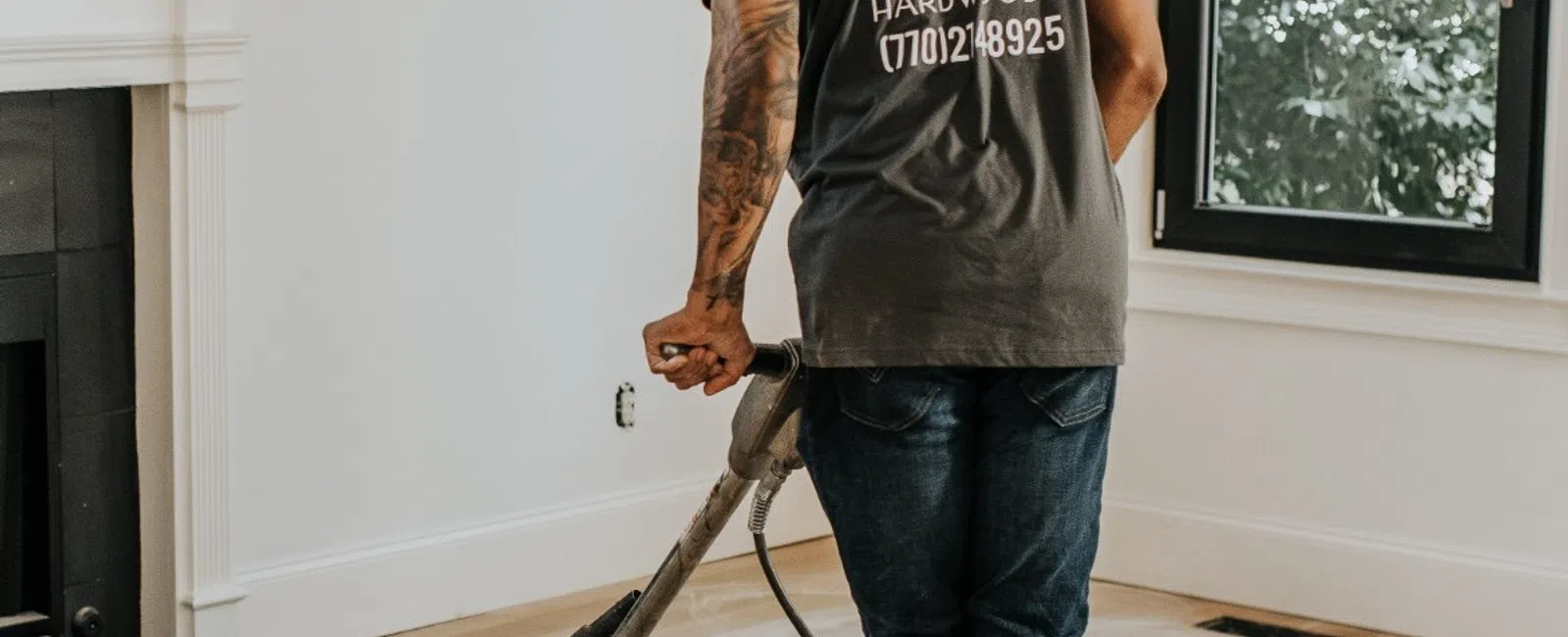 Worker using floor sander to refinish hardwood floors in a bright living room with large windows.
