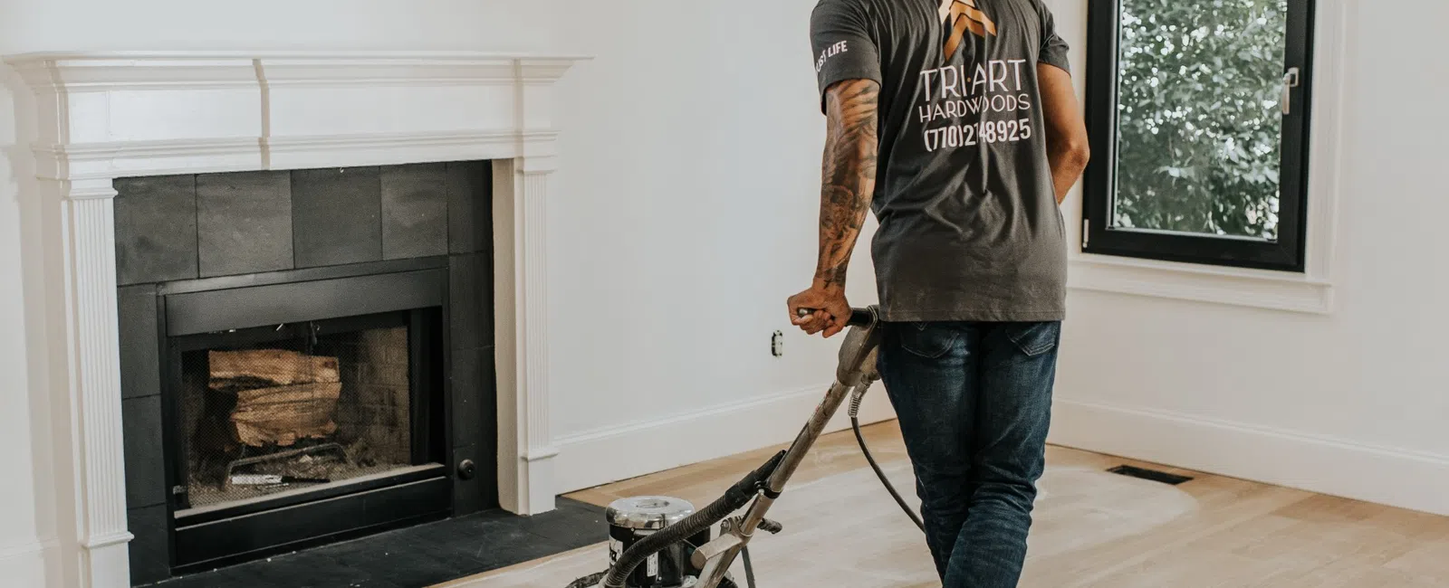 Man sanding hardwood floor near fireplace in bright room with large window and white walls