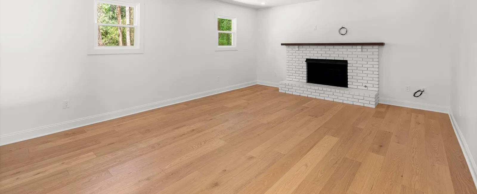 Empty bright living room with light wood flooring, white walls, two windows, and white brick fireplace