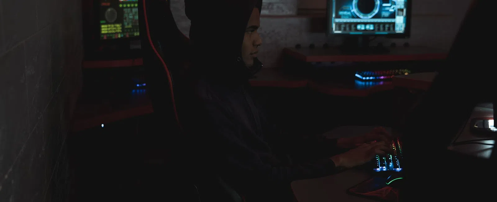 Person in a dark hoodie typing on a keyboard with multiple screens displaying digital graphics in a dim room.