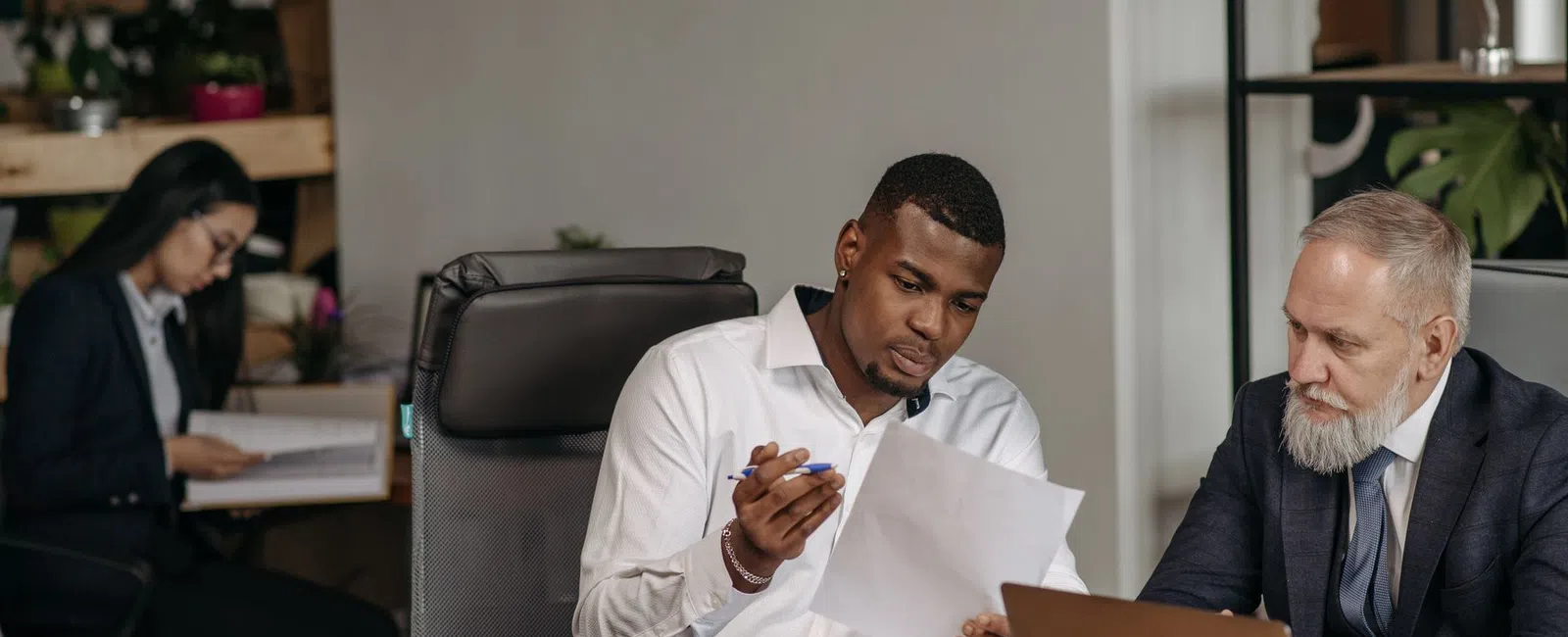 Two businessmen reviewing documents at a desk with laptop while a woman works in the background office