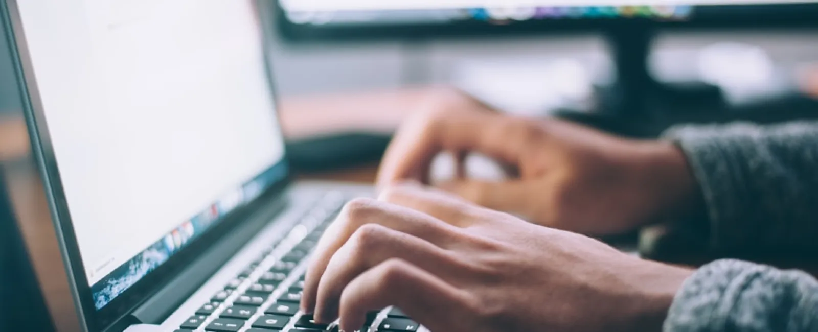 Close-up of hands typing on a laptop keyboard with a blurred monitor in the background in an office setting