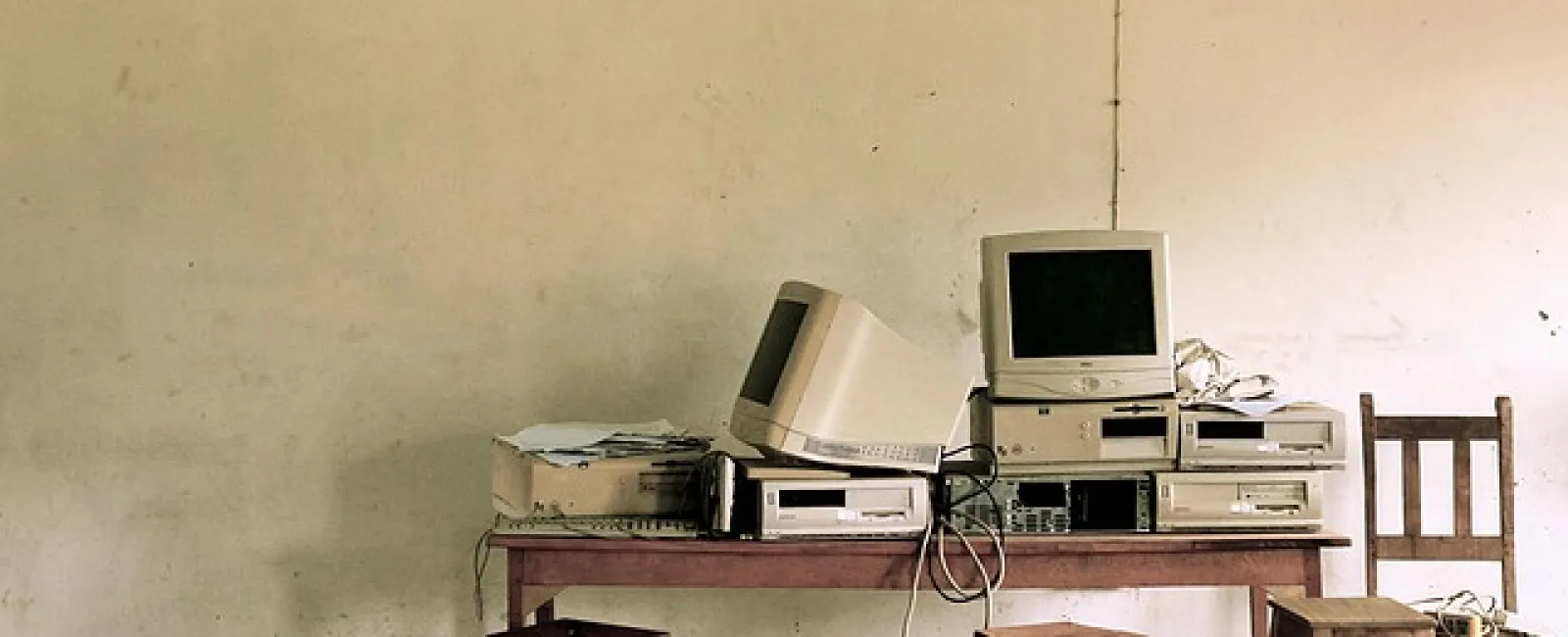 Old computer monitors and CPUs stacked on a wooden table with stools against a plain beige wall.