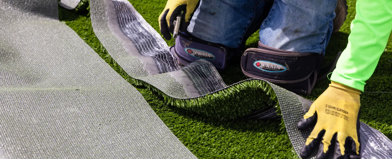 Worker wearing gloves and knee pads installing artificial grass turf outdoors on green surface.