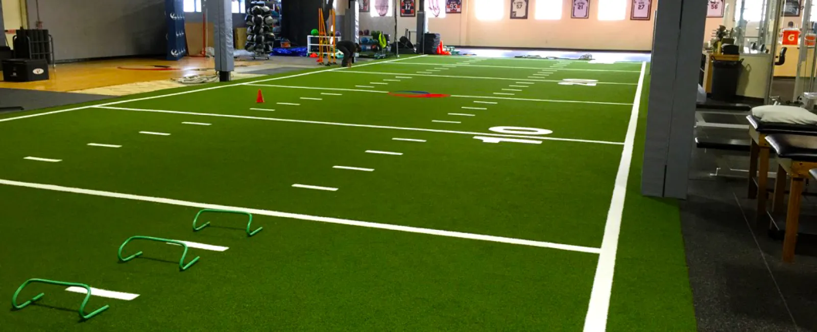 Indoor training facility with artificial turf, agility hurdles, cones, and fitness equipment for sports conditioning.