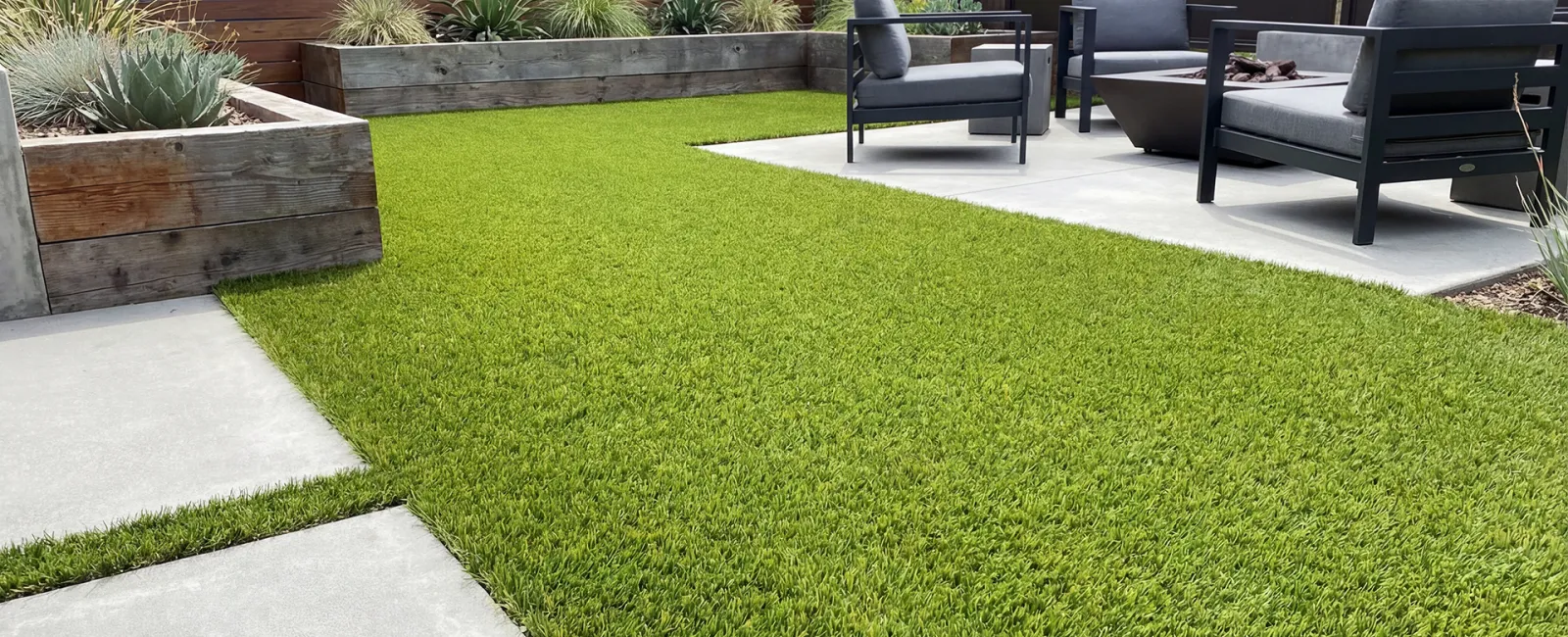 Modern backyard with artificial grass, concrete patio, wooden planters, and black outdoor seating with cushions.