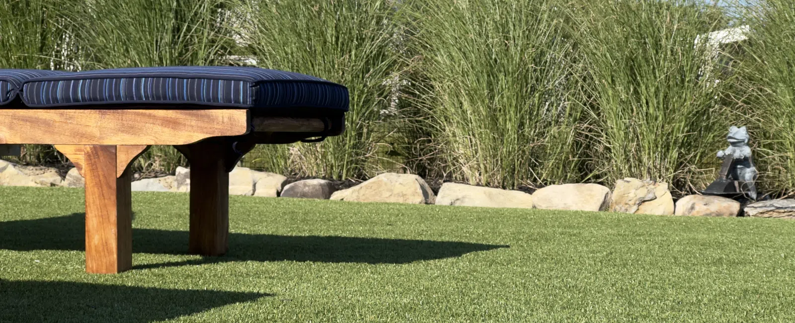Wooden outdoor bench with blue cushion on green grass near ornamental rocks and tall grasses in sunlight
