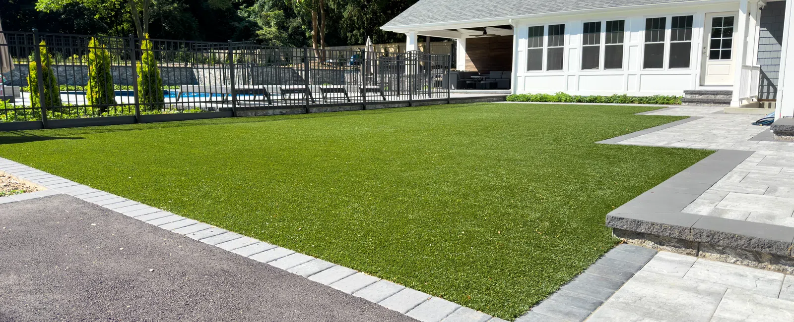 Modern backyard with artificial grass lawn, stone patio, and a pool surrounded by fence on a sunny day.
