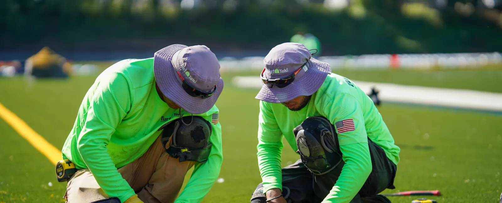 Two workers in bright green shirts and hats installing artificial turf on a sunny day outdoors.