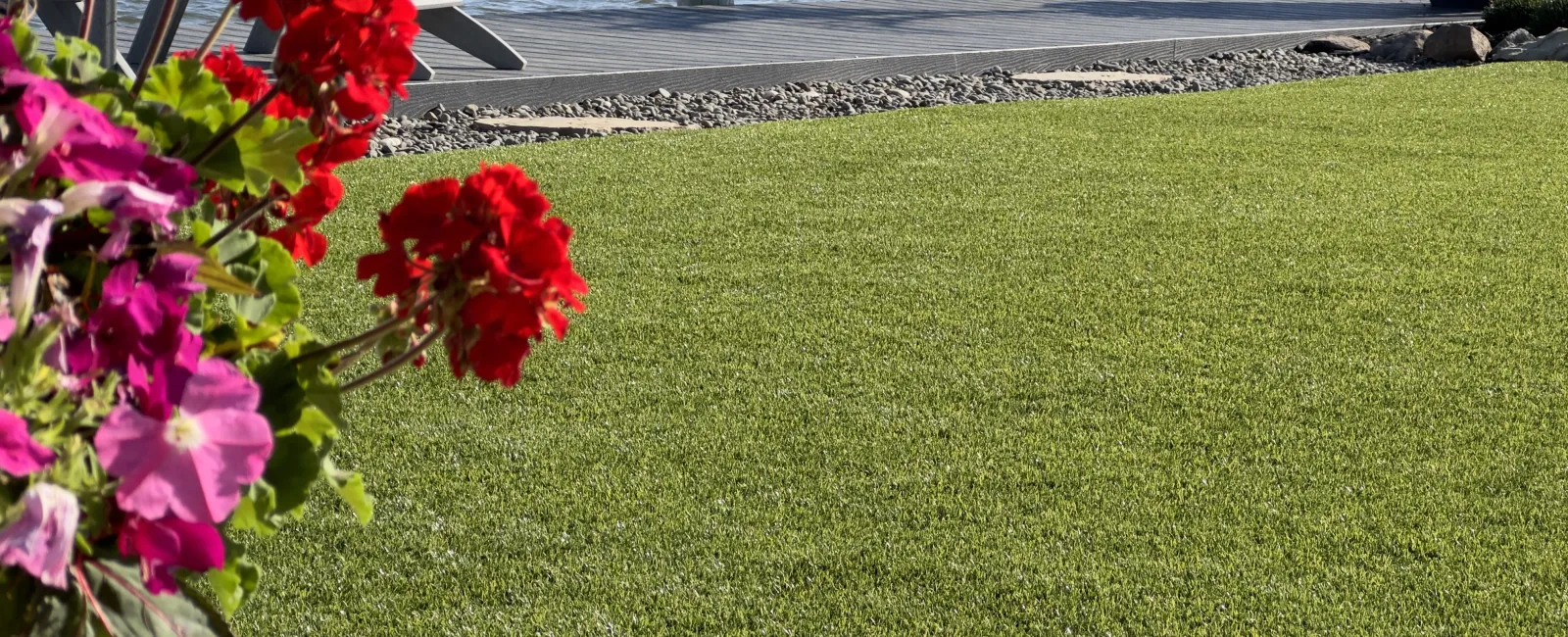 Green lawn with bright red and pink flowers near a wooden dock by calm water on a sunny day