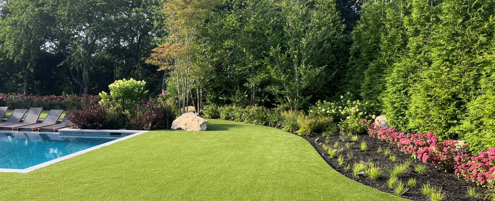Well-manicured lawn beside a swimming pool with lounge chairs, flower beds, and dense green trees under blue sky.