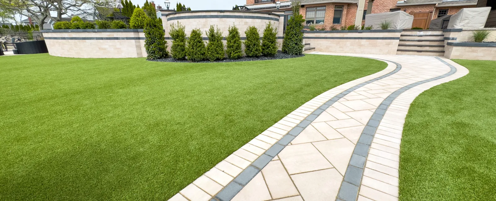 Curved stone pathway through well-manicured green lawn leading to a brick house with garden and shrubs.
