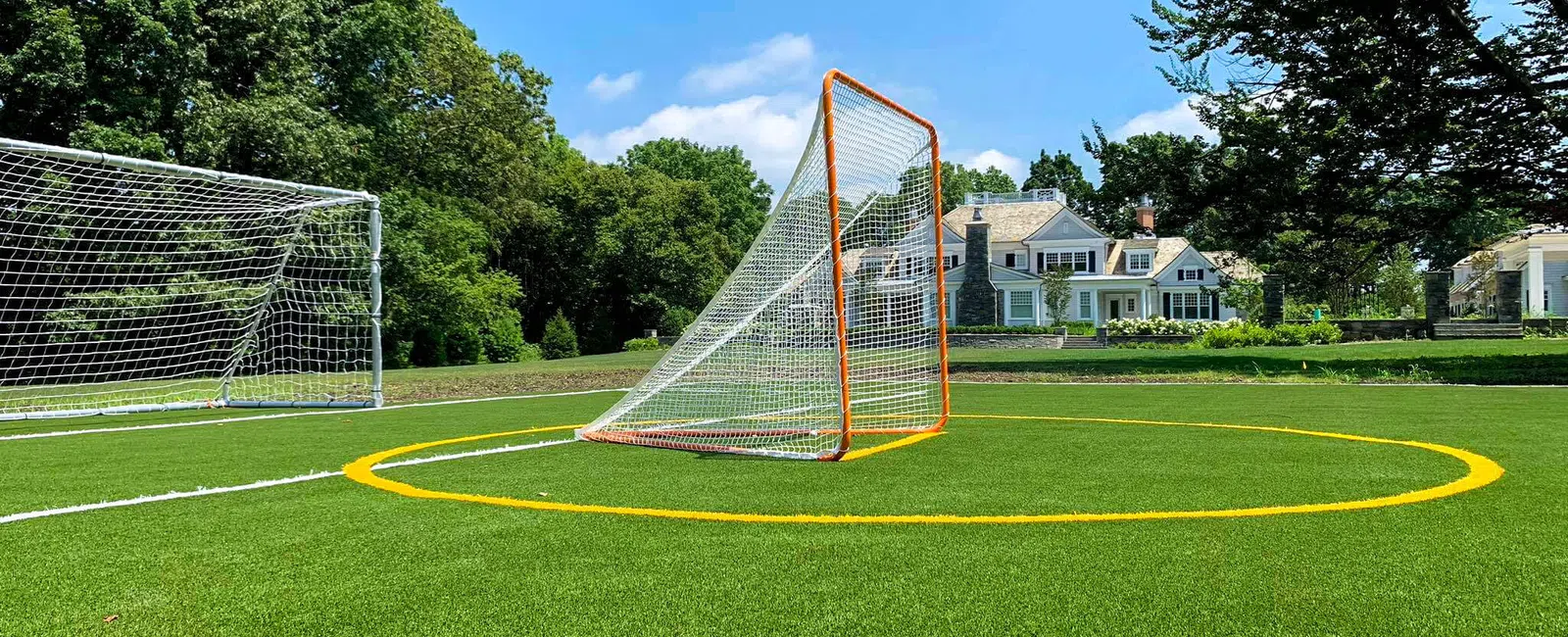 Lacrosse goal and soccer net on green turf field with houses and trees under blue sky