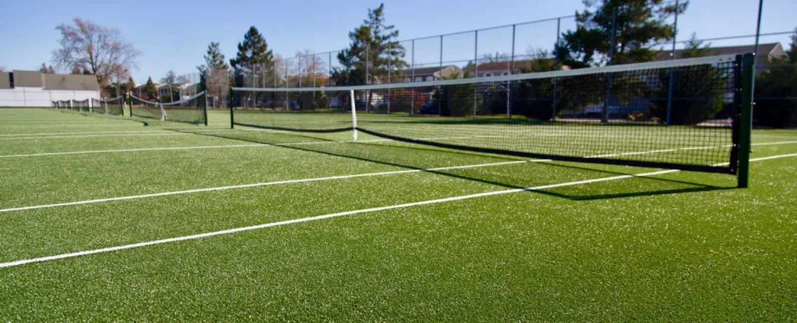 Sunny outdoor tennis courts with green artificial turf, white lines, and black nets under a clear blue sky.