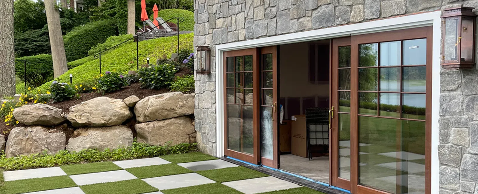 Stone patio with checkerboard pattern of grass and concrete slabs outside a stone house with large wooden sliding doors.