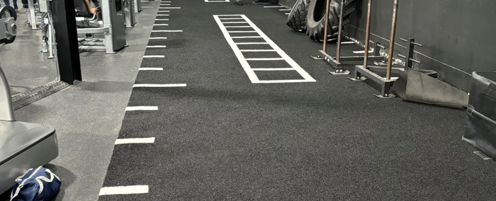 Indoor gym training area with turf, agility ladder markings, large tires, and weight sleds for functional workouts.