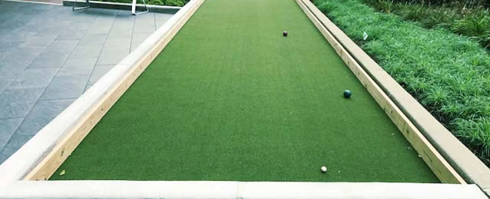 Outdoor bocce ball court with green turf, three bocce balls, surrounded by plants and patio furniture.