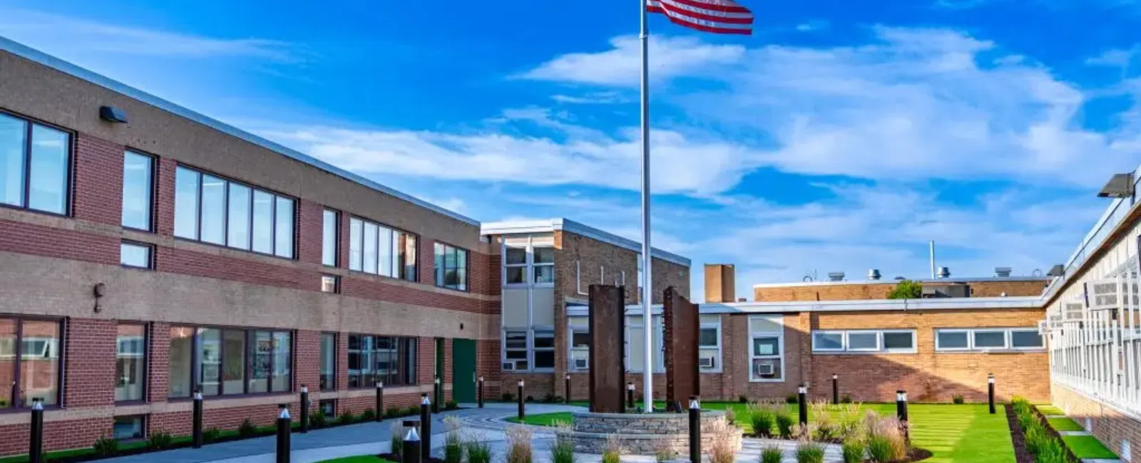 American flag flying in front of a modern school building with a curved walkway and green lawn under a blue sky