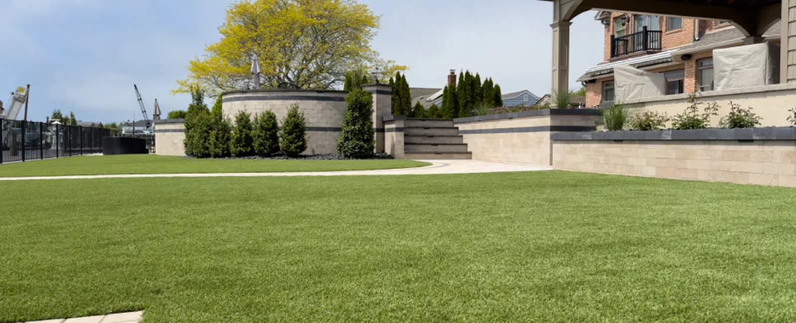 Well-maintained green lawn with stone retaining walls, shrubs, patio, and a pergola-covered seating area on a sunny day.