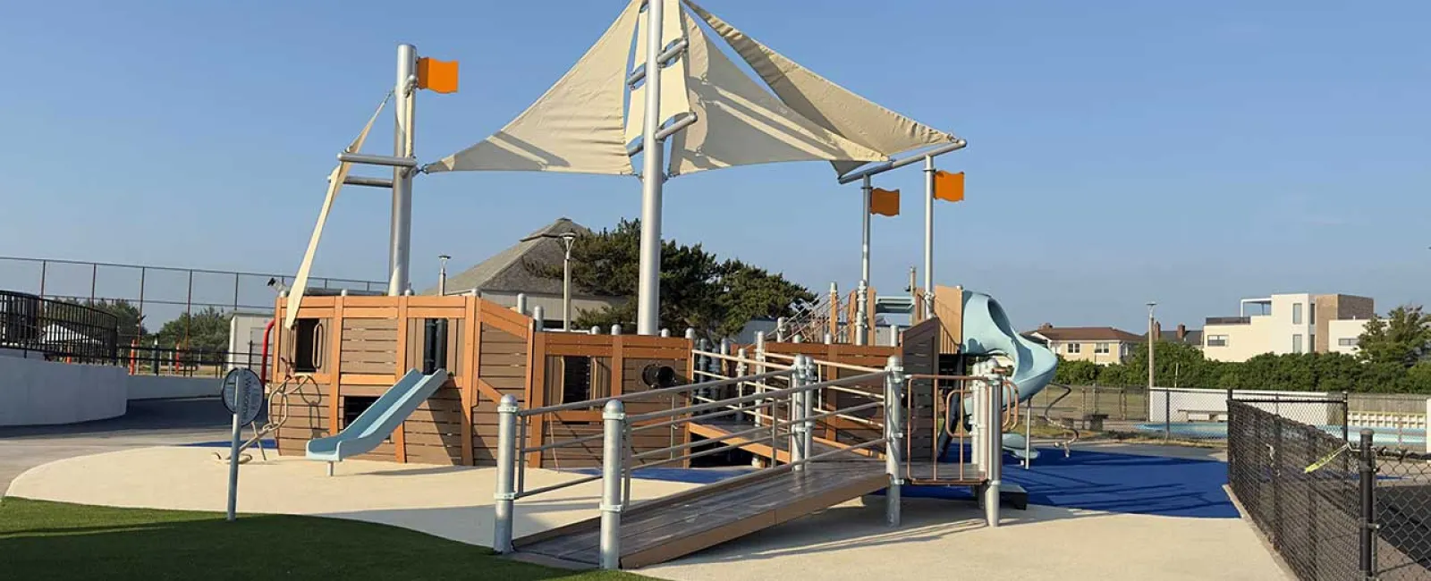 Modern pirate ship-themed playground with slides, ramps, orange flags, and shaded sails under a clear blue sky.