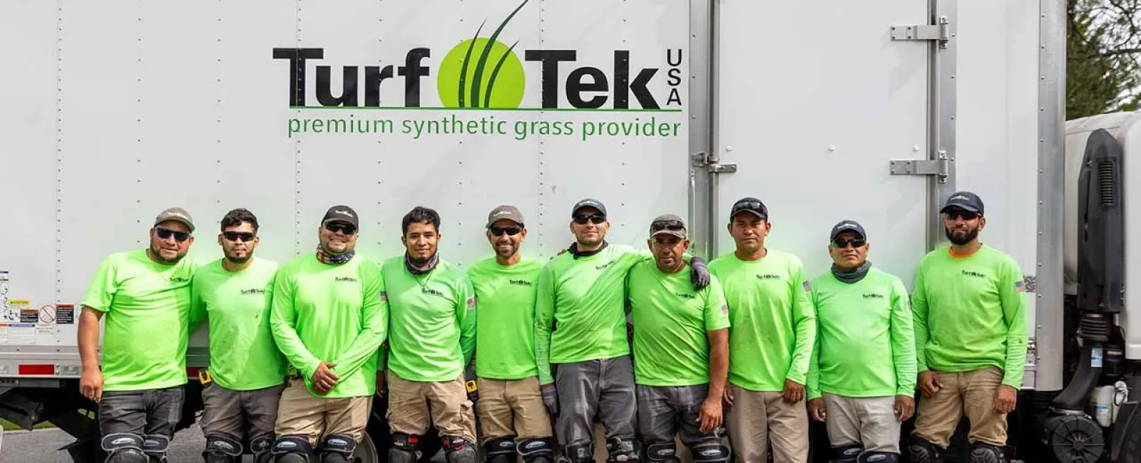 Team of workers in green shirts and knee pads standing in front of Turf Tek USA truck with company logo.