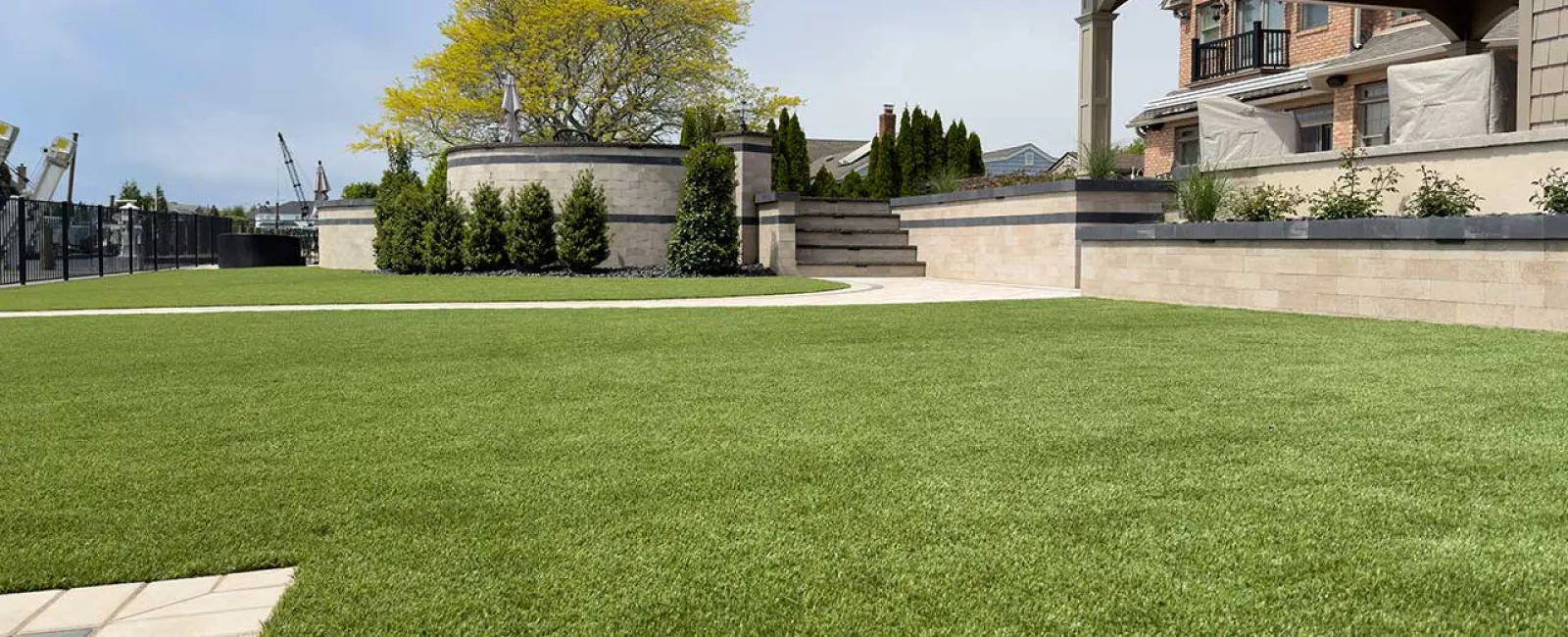 Well-maintained artificial lawn with stone retaining walls, shrubs, a tree, and a covered patio seating area.